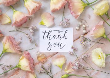 Card with the handwritten text THANK YOU surrounded by pink flowers, earrings, petals and parfume flacon around top view on a marble table. Romantic concept